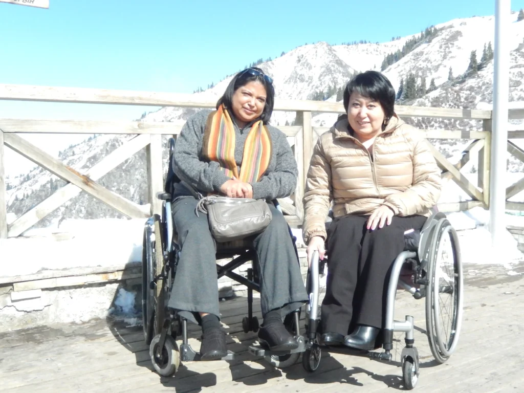Two women in wheelchairs smiling in front of a snowy mountain backdrop.