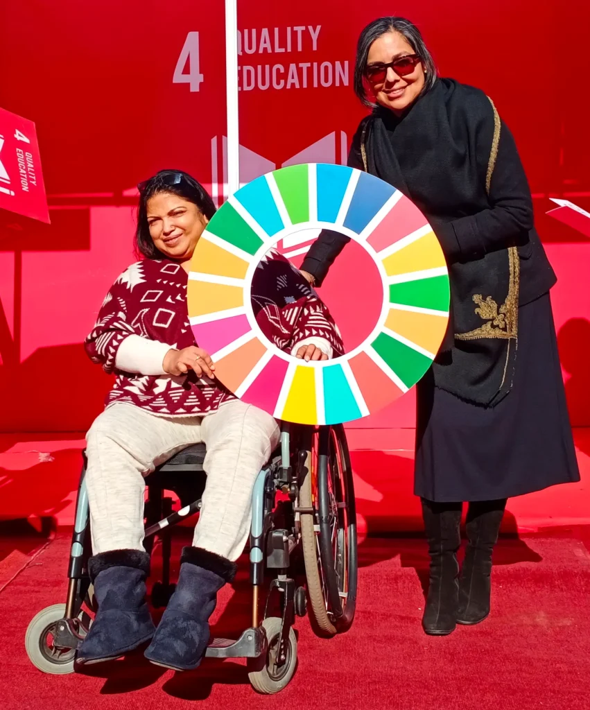 Two women, one in a wheelchair, holding a colorful circular graphic of the Sustainable Development Goals (SDGs) in front of a red banner that reads "QUALITY EDUCATION."