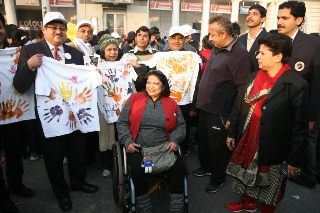 A group of people, including a woman in a wheelchair, are outdoors holding up white t-shirts decorated with colorful handprints.