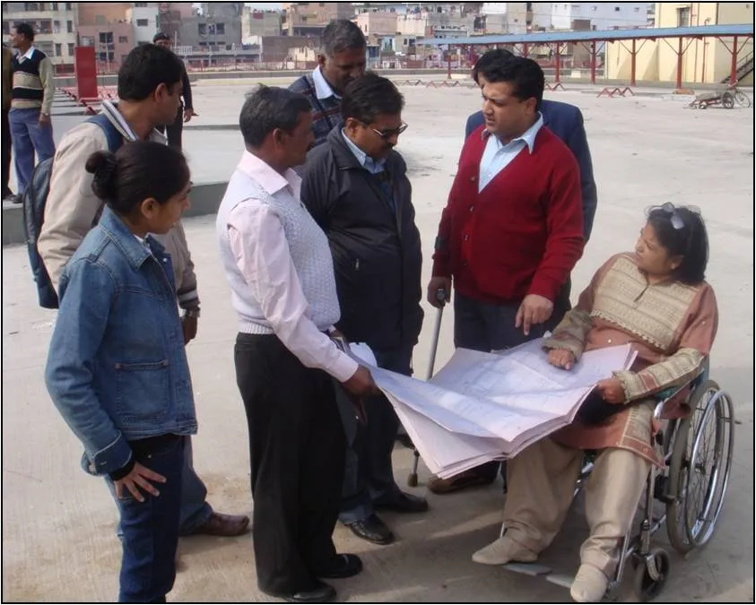 A group of people, including a woman in a wheelchair, reviewing blueprints or plans outdoors on a rooftop.