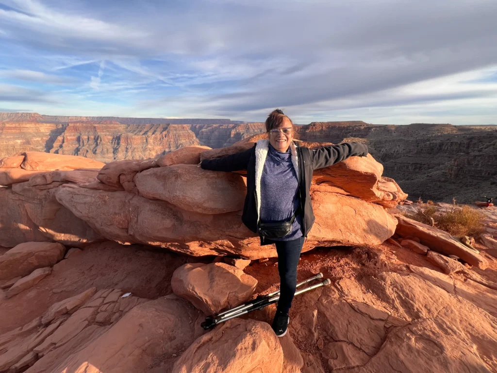 A smiling woman, Veneranda Mateo, stands on a large reddish-brown rock, with her arms outstretched behind her. She is wearing a dark jacket and glasses, and two crutches are resting in front of her. In the background, a vast canyon landscape stretches under a partly cloudy sky.