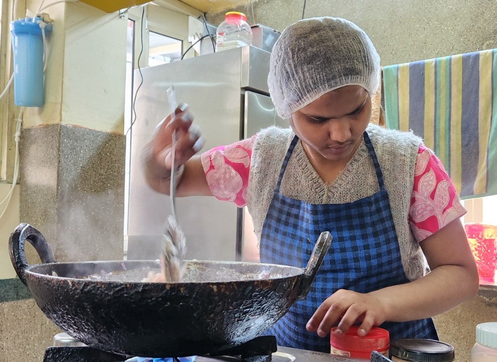 A woman wearing a blue and white checkered apron and a hairnet is stirring food in a large black wok on a gas stove. She is focused on the task, with a red container of spices nearby.