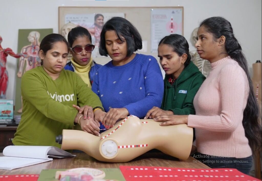 A group of five women stand around a table, focused on a beige anatomical model of a torso. The woman in the center, wearing a blue sweater, and the woman to her left, in a green sweater, are pointing at and touching the model. The other three women are watching intently
