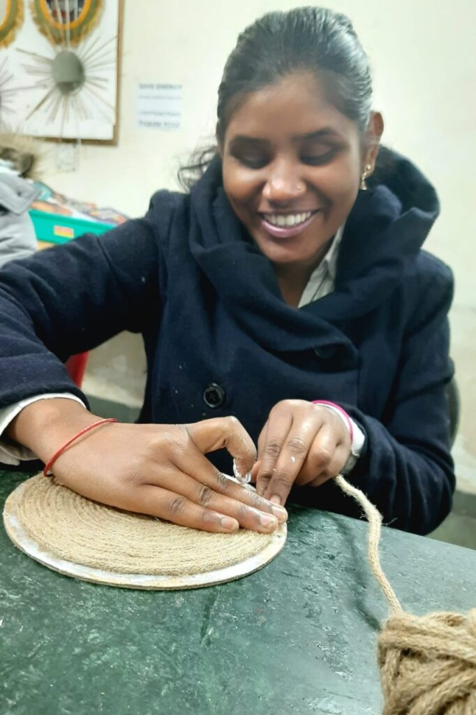 A woman with long dark hair, wearing a black coat, smiles as she crafts with a long piece of thick twine. She is sitting at a table and wrapping the twine around a flat, circular base. A ball of twine is on the table to her right.
