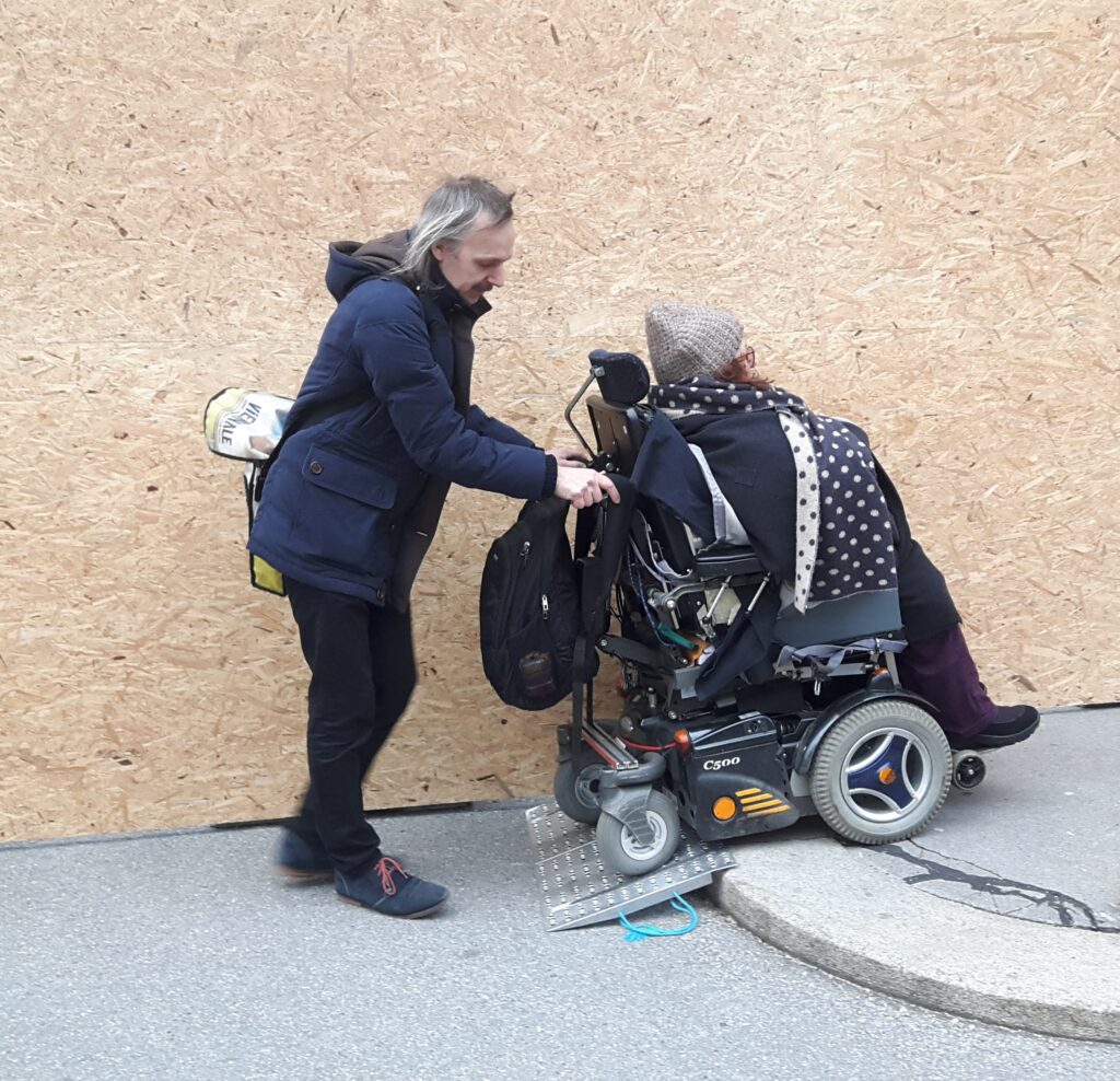 A man in a blue jacket is helping a woman in a motorized wheelchair go up a small ramp onto a sidewalk. The woman is wearing a dark coat and a patterned scarf. The background is a plain wooden wall.
