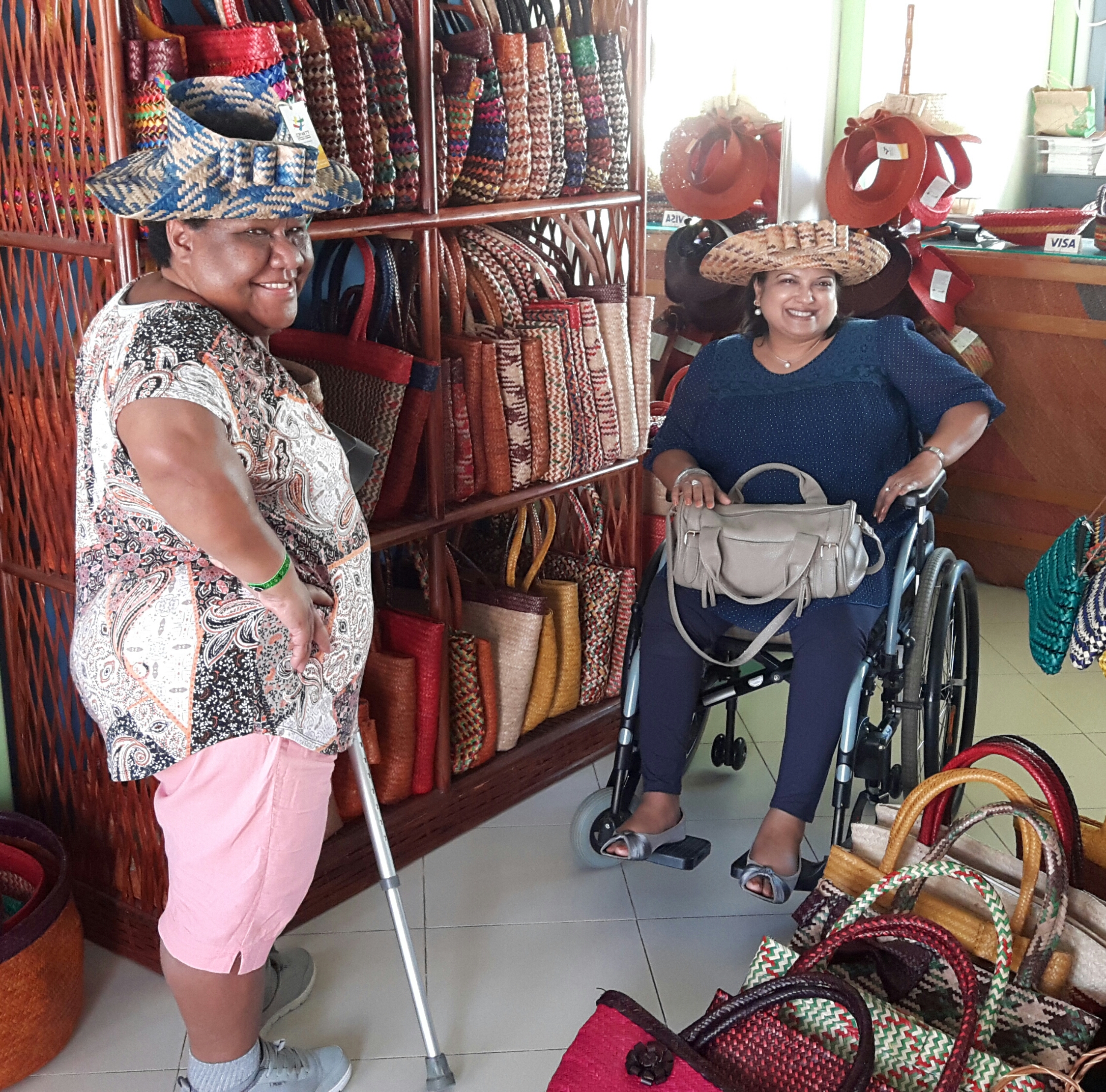 Two women, one standing with a walking stick and the other in a wheelchair, pose for a photo inside a shop. Both are wearing large straw hats and smiling. The shop is filled with woven bags and hats.