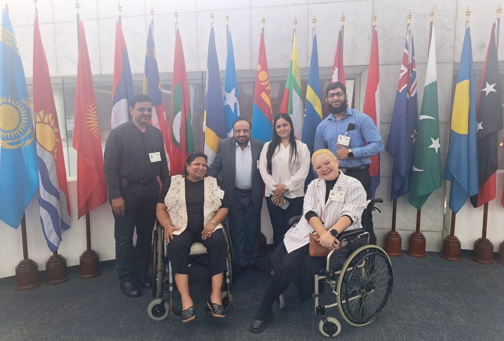 This image shows 4 people standing and two women in a wheelchair, who are happily posing for a picture in front of flags from different countries.