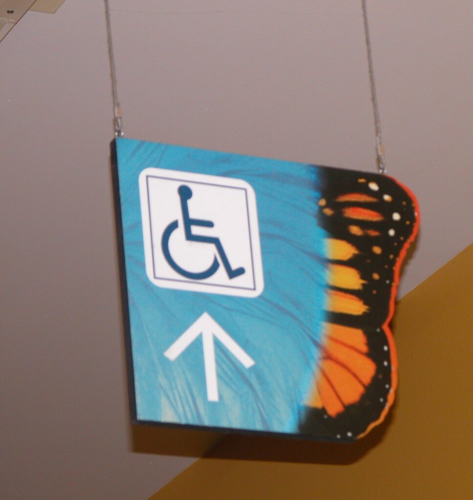 A square sign with a monarch butterfly wing on the right side. The sign has a blue background with a white wheelchair accessibility symbol and a white arrow pointing up. The sign is hanging from a ceiling by two thin wires.