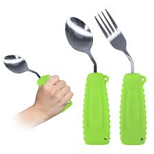 Three eating utensils with thick, green ergonomic handles are shown on a white background. One is a spoon being held by a hand. The other two, a fork and a larger spoon, are shown from the side, demonstrating their design. The utensils have bent heads to make them easier to use.