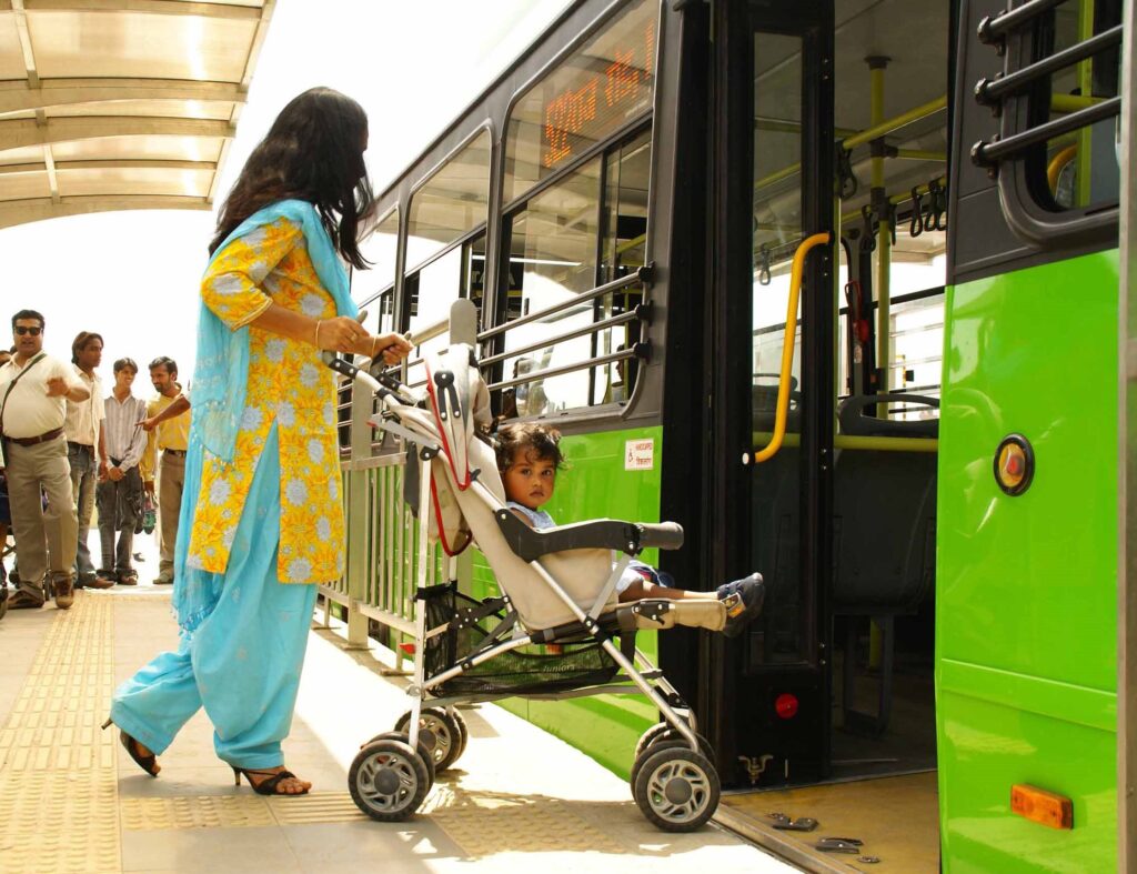 A woman in a blue and yellow patterned tunic and a young child in a stroller are waiting to board a green public bus. The bus is at a stop with a curb ramp, and other people are standing behind them.
