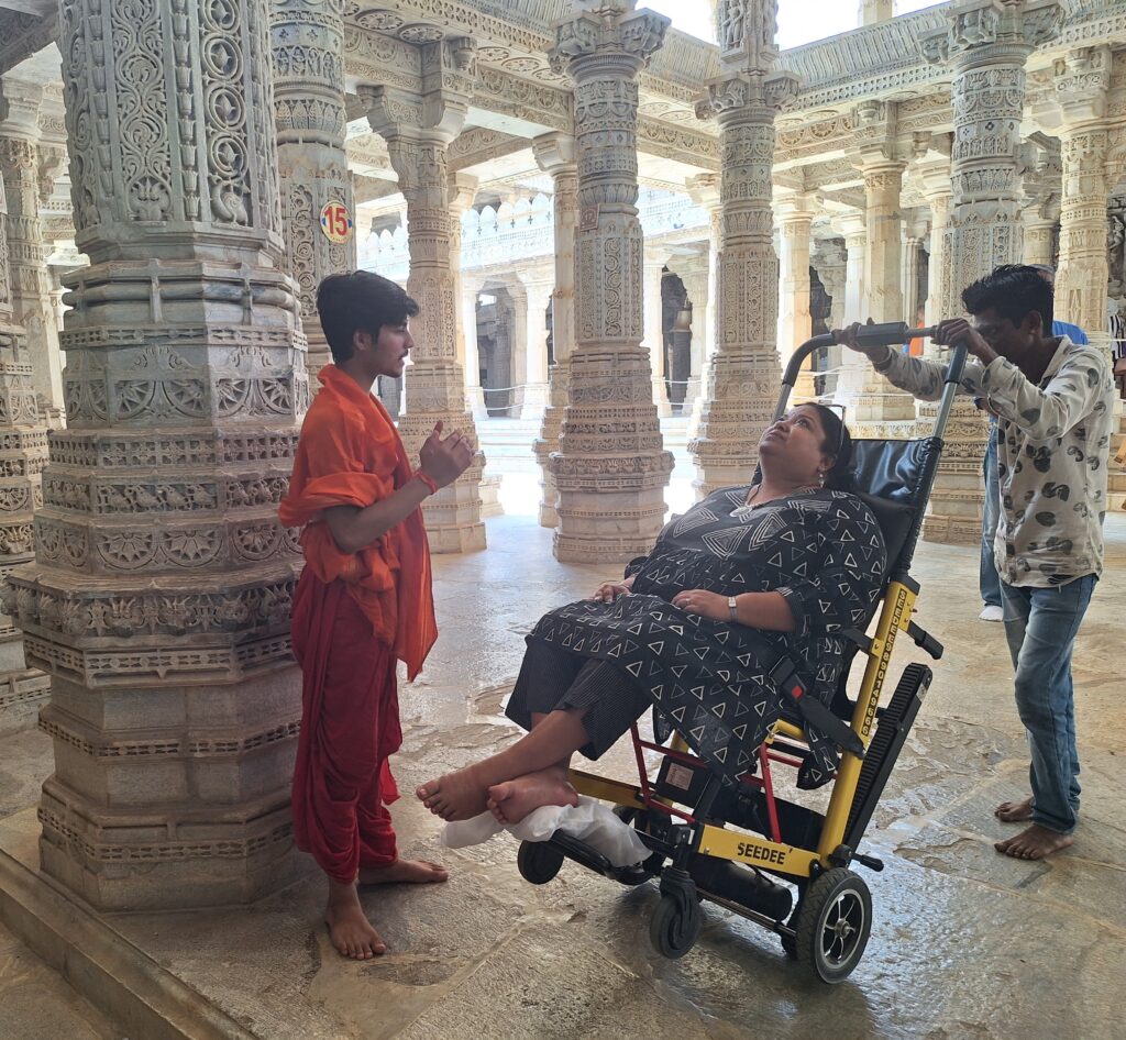 A woman in a yellow wheelchair is being pushed by a man inside a temple. A young boy in an orange robe is standing in front of them, praying.
