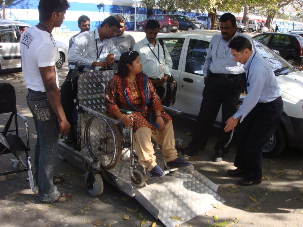A group of people are gathered outdoors, with a woman in a wheelchair on a portable ramp attached to a small cart. Several men are standing around her, looking at the device.