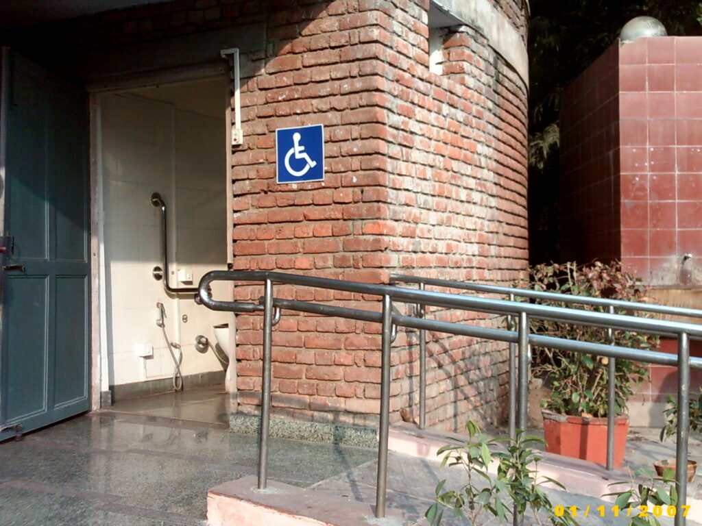 n accessible public restroom with a brick exterior. A blue wheelchair accessibility sign is mounted on the wall. A metal ramp with a handrail leads to the entrance. Inside the open door, a grab bar is visible next to a toilet