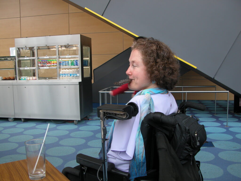 A woman in a wheelchair, with curly brown hair, sits at a wooden table in an indoor space.