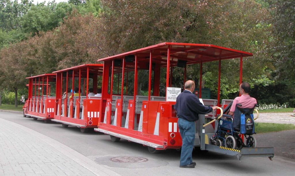 The image shows a red toy train where in a person in a wheelchair is sitting at the end with another person standing next to him.