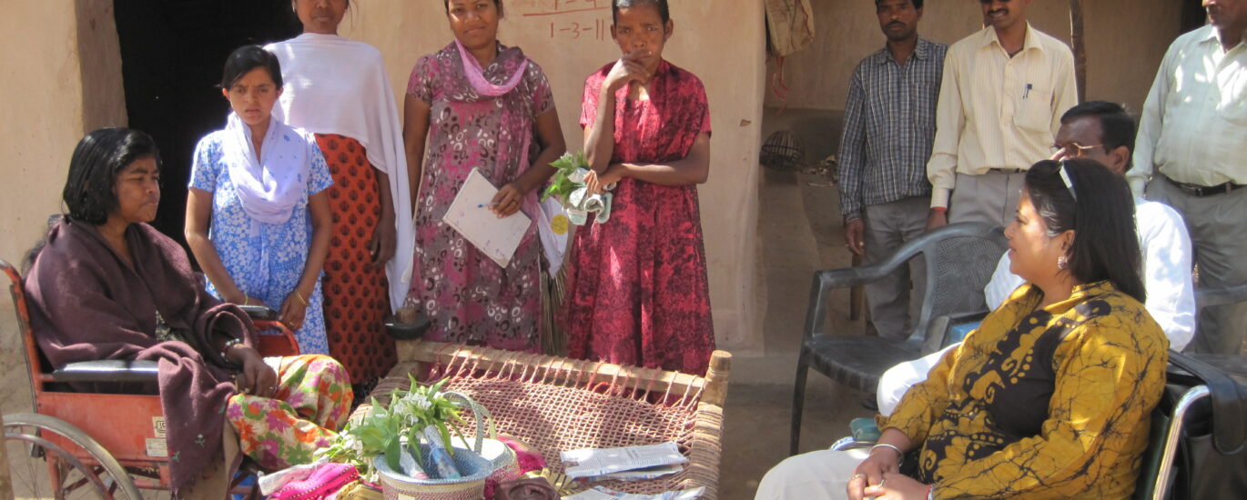 A picture where there are people gather around a hut in a village and two women are in a wheelchair having a conversation.