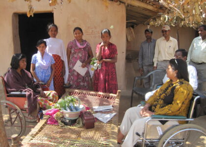 A picture where there are people gather around a hut in a village and two women are in a wheelchair having a conversation.