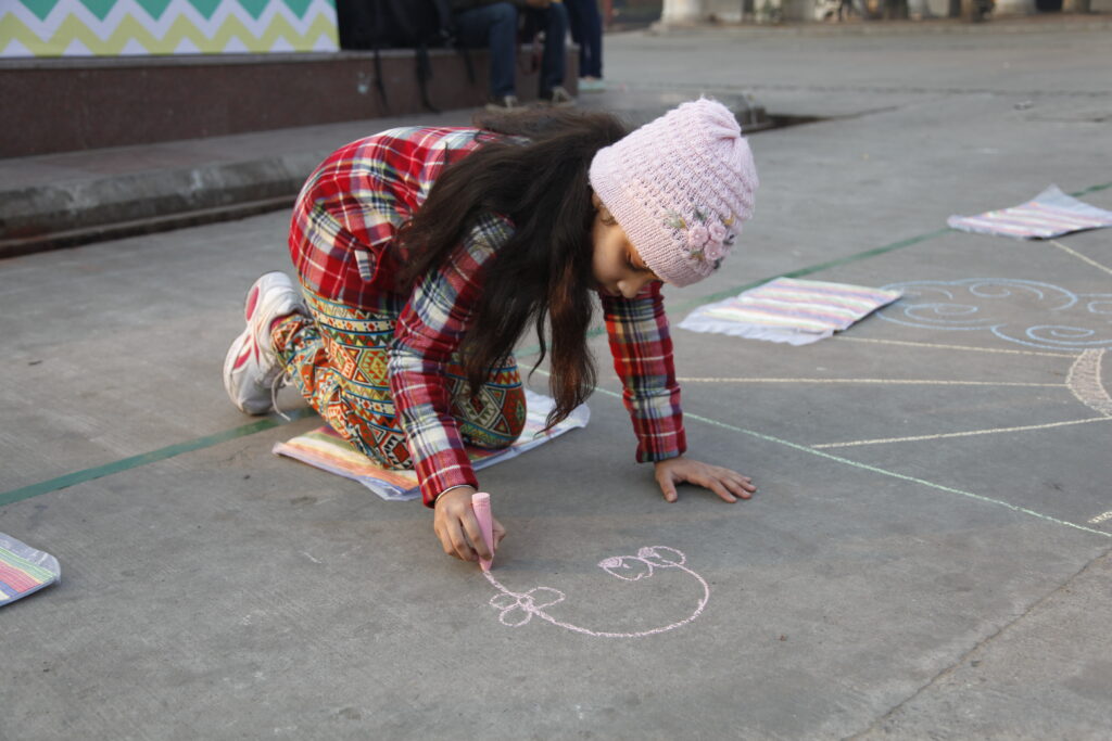 A young girl in a plaid shirt and a pink beanie kneels on a concrete pavement, drawing a flower with pink chalk. A small bag and other drawings on paper and the pavement are visible around her.