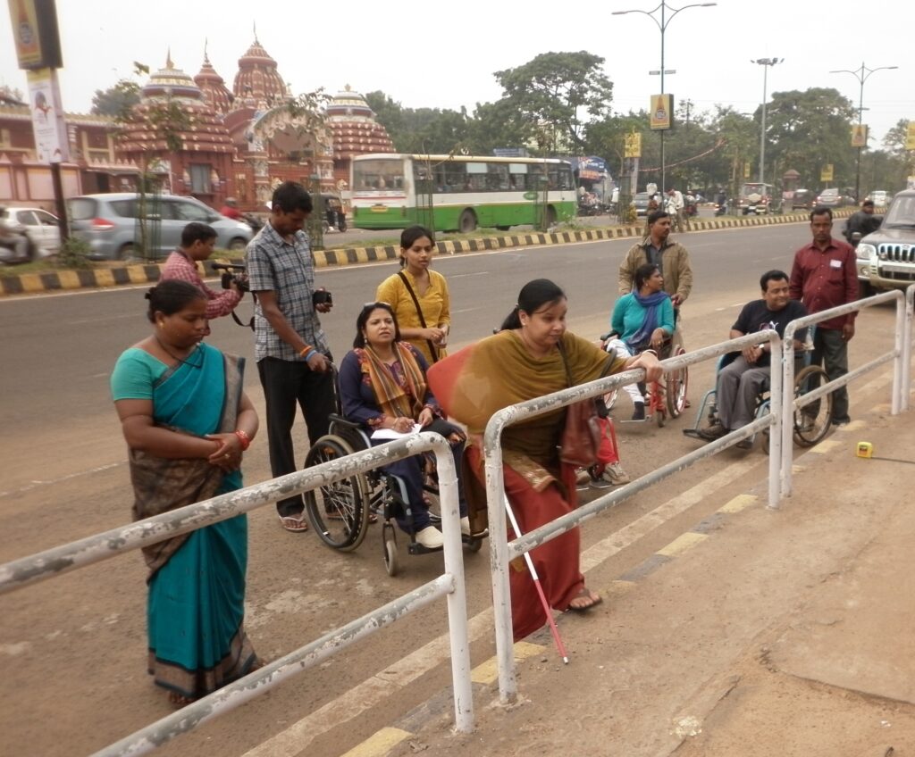 A group of people, some in wheelchairs, are gathered on the side of a road with a metal barrier. They are standing or sitting near a busy street with cars and a bus, and distinctive temple-like buildings are visible in the background. The weather is overcast.