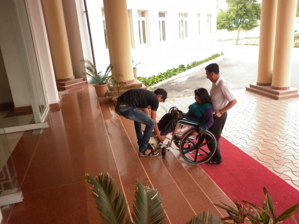 A group of three people, including a woman in a wheelchair on a red ramp, are at the base of stairs leading to a building. Two men are assisting her, one bent down near the wheelchair and the other holding the handles.