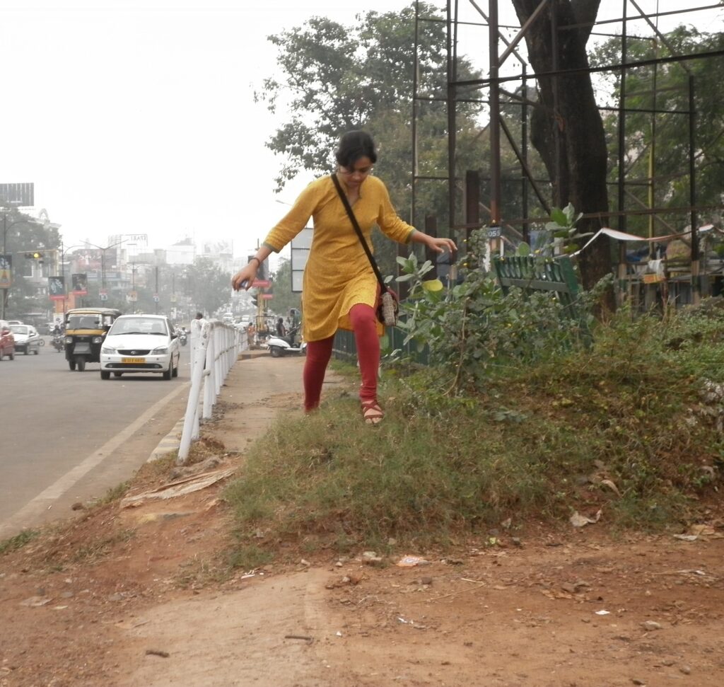 A woman in a yellow kurti and red leggings is walking down a grassy slope next to a road with a white guardrail. Cars and a yellow auto-rickshaw are visible in the background, along with buildings, trees, and utility poles. It is an overcast day.