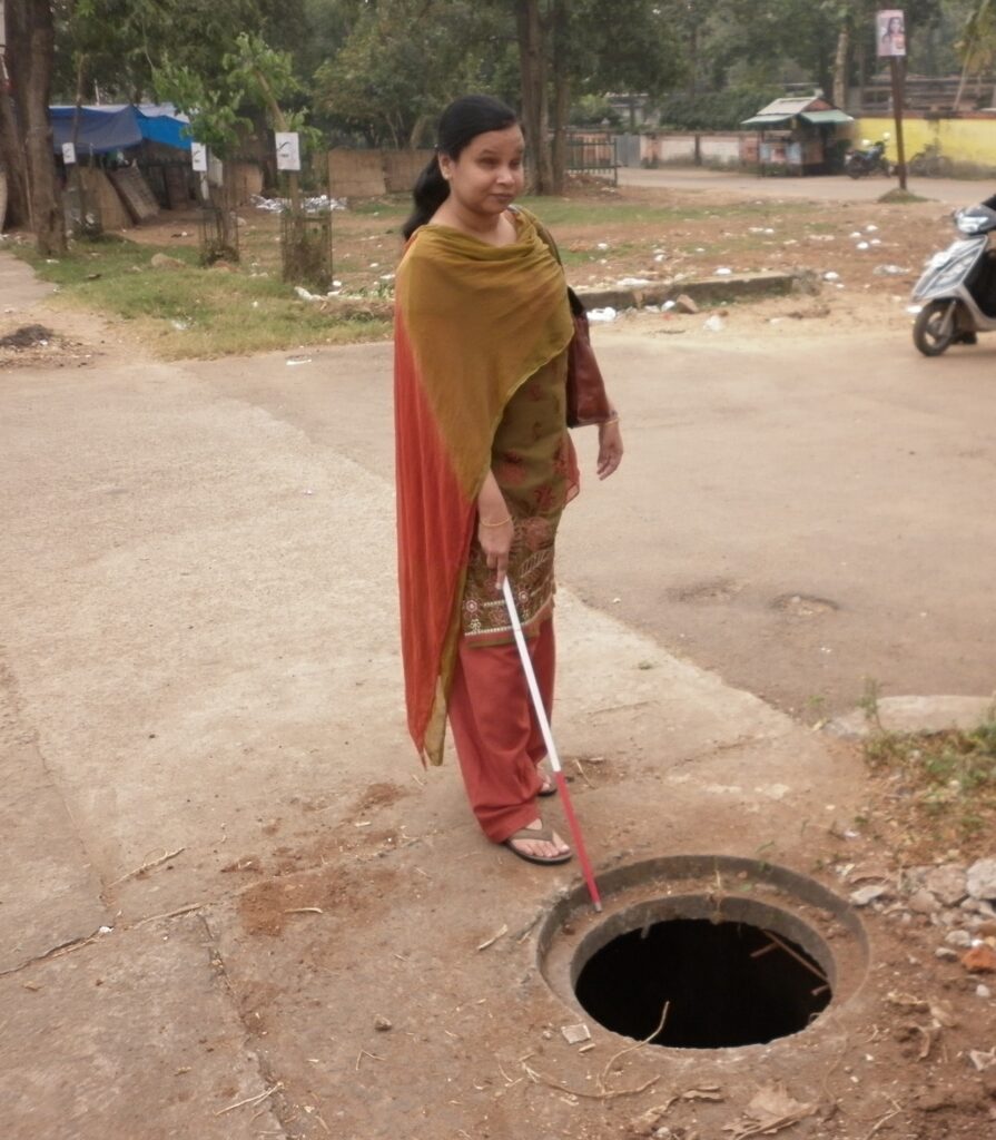 A woman with a white cane is standing by an open manhole on a dirt path.