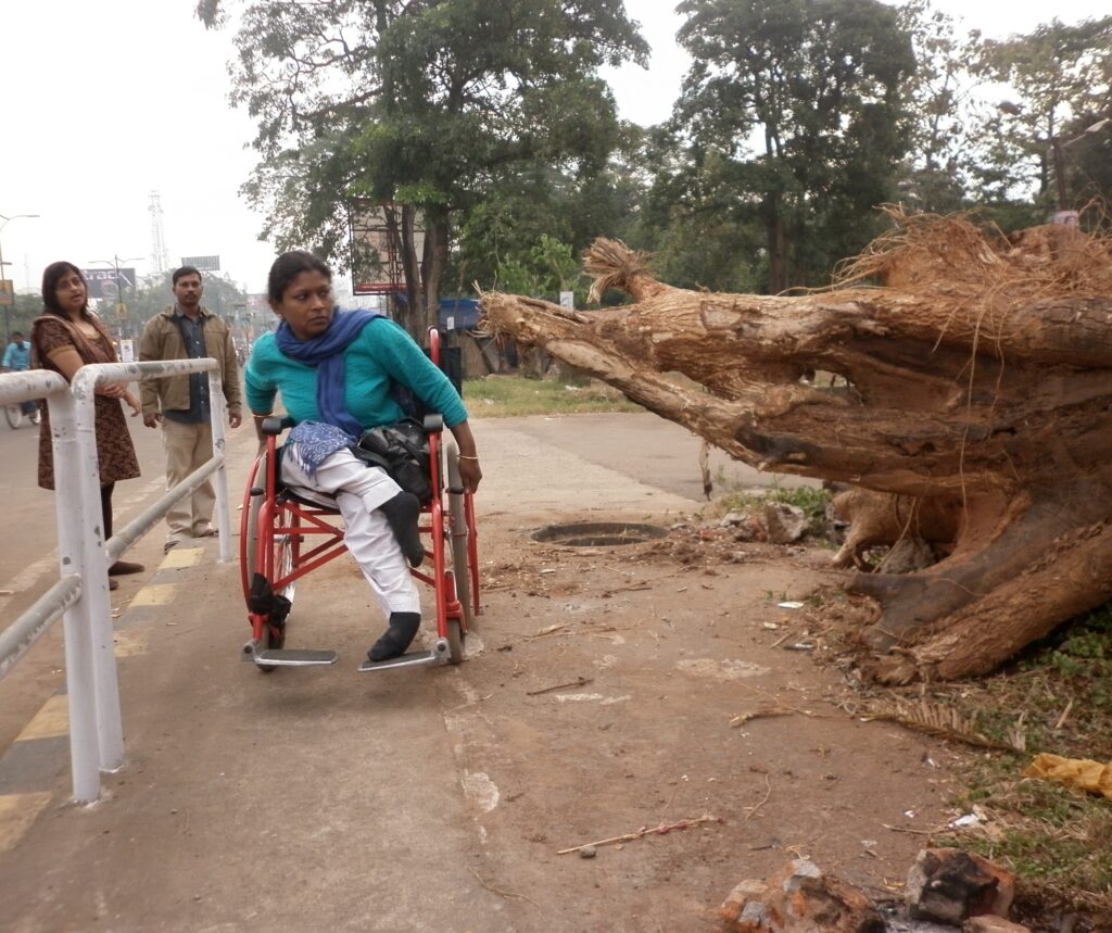 A woman in a red wheelchair, wearing a turquoise top and white pants, is navigating a paved area next to a large, uprooted tree trunk. She is looking to her left, seemingly at the challenge of the uneven terrain.