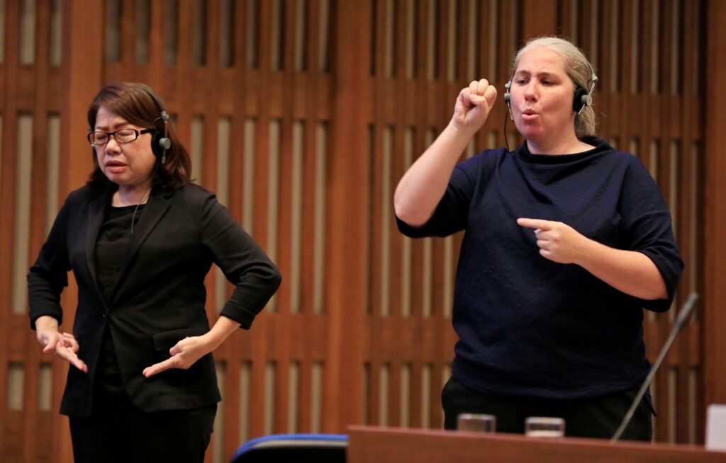A picture of two woman, where both the woman are using sign language to communicate and are wearing headphone.