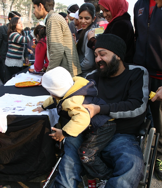 A man in a wheelchair is holding a child on his lap while they participate in a craft activity at a table.