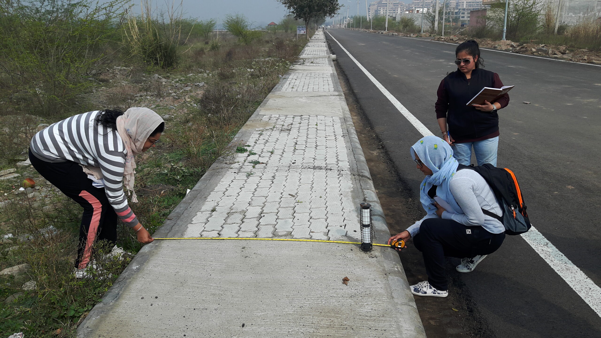Three women measuring the width of a sidewalk next to a road, one with a measuring tape and the other two observing and taking notes.