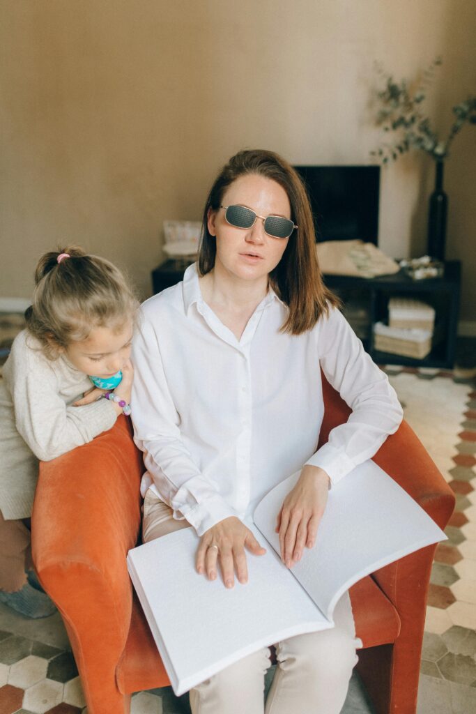 The image shows a woman sitting in a chair, wearing glasses and reading using her hands. A small girl is standing next to the woman.