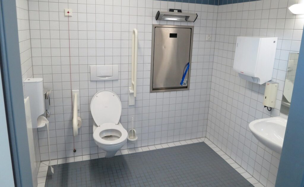 The image shows an accessible public toilet designed for people with disabilities. It features a toilet with a white grab bar on the left side and another one that can be raised or lowered on the right side